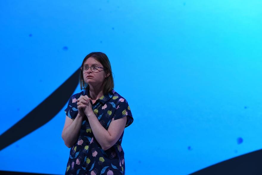 A woman with Down Syndrome stands with her hands clasped. The wall behind her is lit blue.