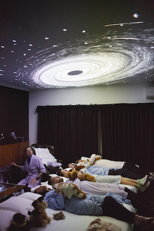 four people lying on hotel beds, in dressing gowns, holding teddies and watching projections of a galaxy on the ceiling