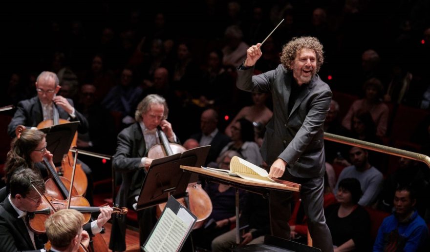 A smiling conductor directs the orchestra from the podium