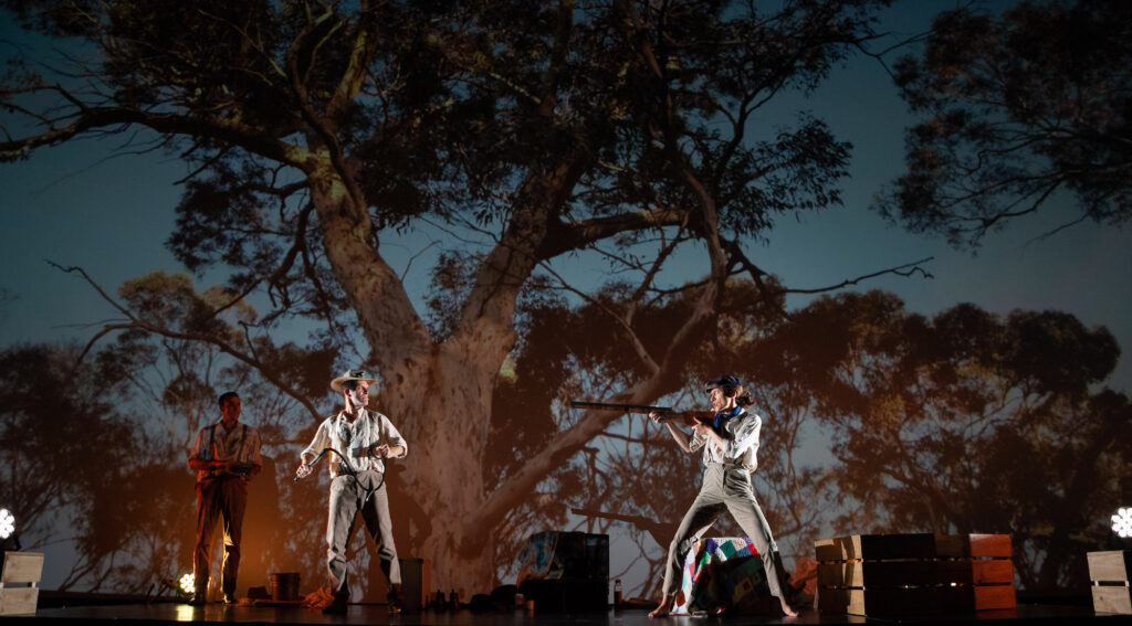 A woman dressed as a man holds a large gun up to two men. They are dressed in trousers, shirts and braces that suggest a scene from the early 20th century. The backdrop is of West Australian bush; a huge gum tree is in the foreground.