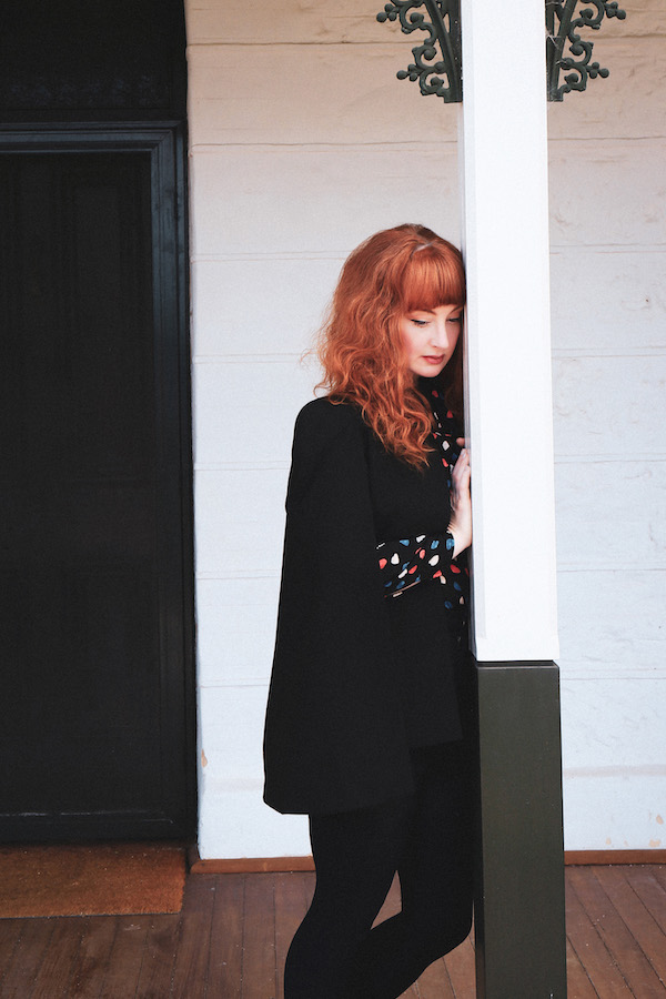 Rachael Dease leans against a wooden post, her eyes cast down. Her red hair is offset by the white of the building behind her and her black outfit with splashes of red, blue and white at collar and sleeves.