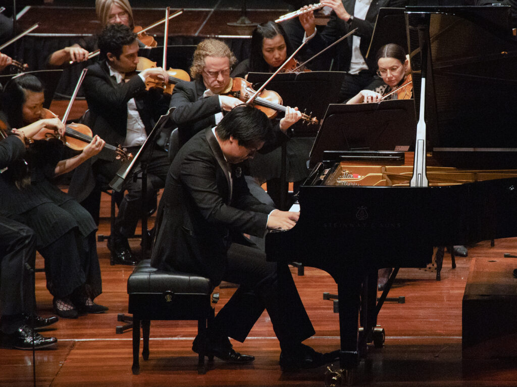 A man in a suit crouches over a piano keyboard, behind him are orchestral musicians