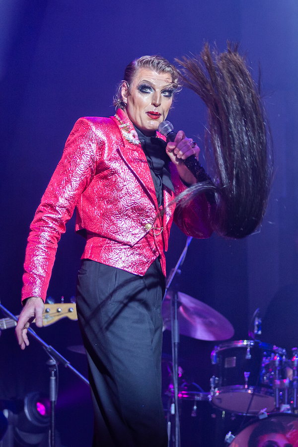 Reuben Kaye stands holding a microphone with a long artificial pony tail. He stands at right angles, with his head turned to the camera, and appears to be mid-sing or speech. He wears a shiny hot pink jacket and black dress pants.