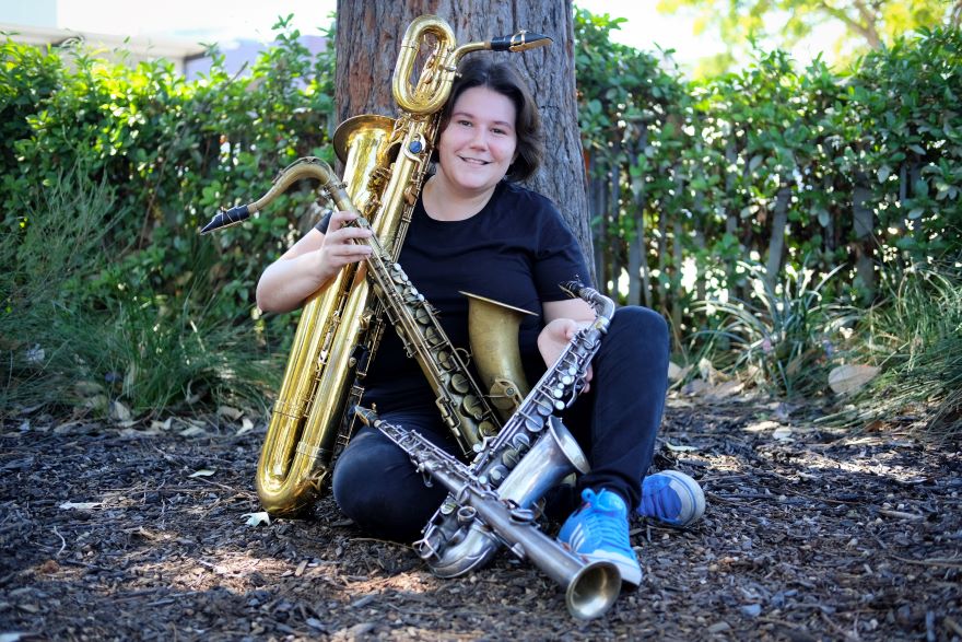 A woman leans against a tree holding four saxophones in her lap
