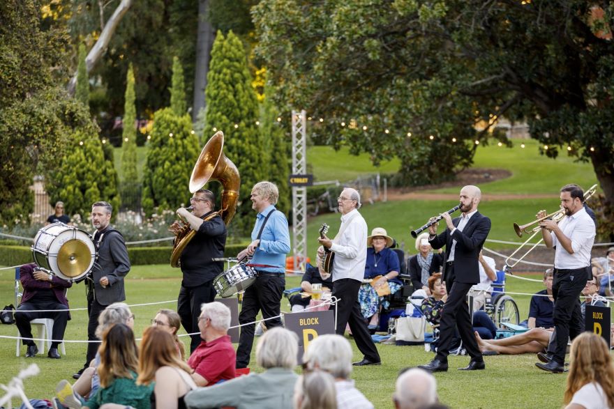 A line of musicians walk along the grass, playing their instruments while they are watched by picnickers