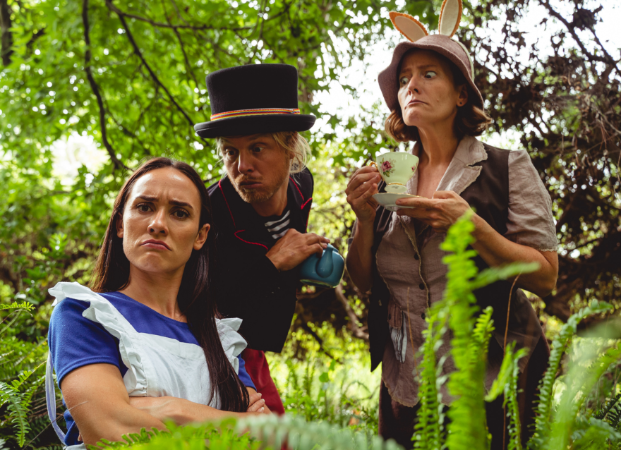Three people crouch among some ferns: a woman in a blue dress frowns, a man with a top hat leans in and a woman with hare ears on her hat sips from a tea cup