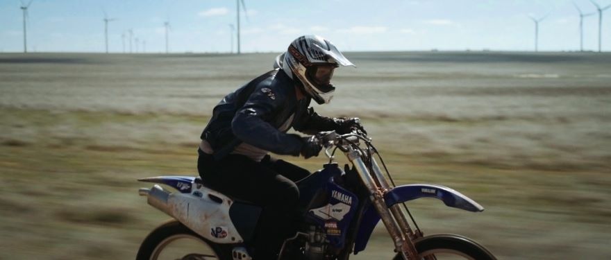 A man crouches over a motorbike , behind him in the paddock are wind turbines