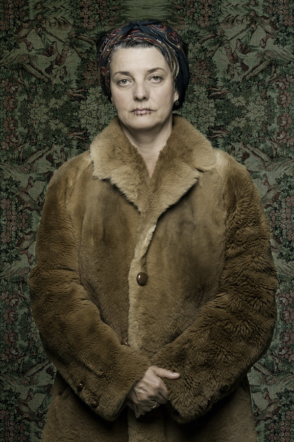 A photograph of Olga Cironis with her mouth stitched shut. She wears a fur coat and a dark green patterned scarf. She stands against wallpaper patterned with forest-like images.