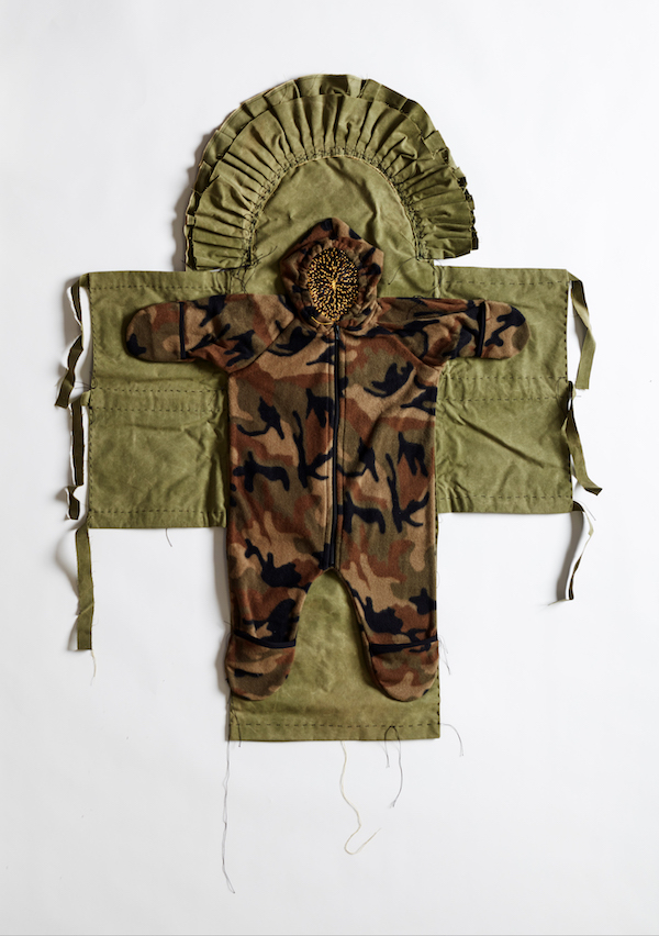 A baby's romper, made from camouglafe material, is shaped onto a crucifix shaped piece of khaki material, with a frill around the head.