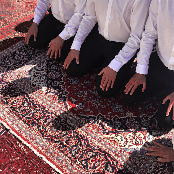 Three men kneel on a Persian carpet, their hands on their knees. They are dressed in white shirts and black pants. Their heads and shoulders are cropped out of the frame.