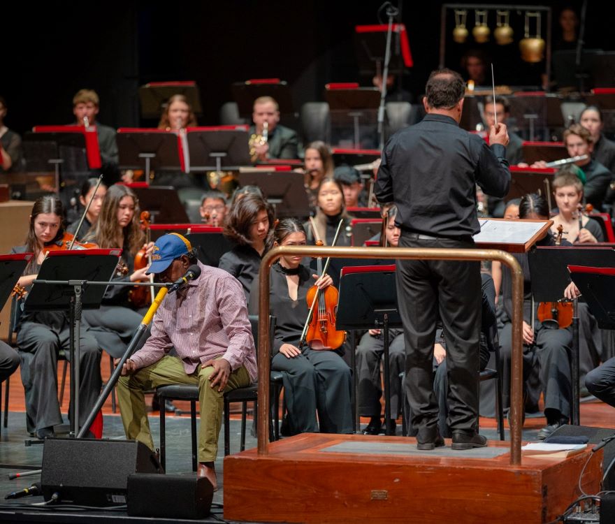 A man wearing a cap plays the lambirlpil (didgeridoo) in front of a youth orchestra, with a conductor on the podium.
