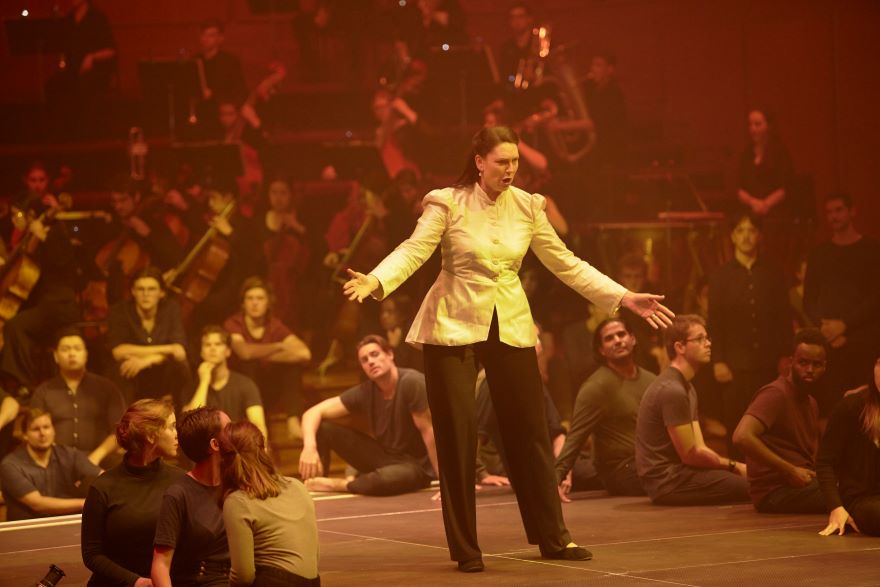 A woman in a white jacket sings with arms outstretched, bathed in hazy orange light. She shares the stage with people in casual clothes, seated on the floor.