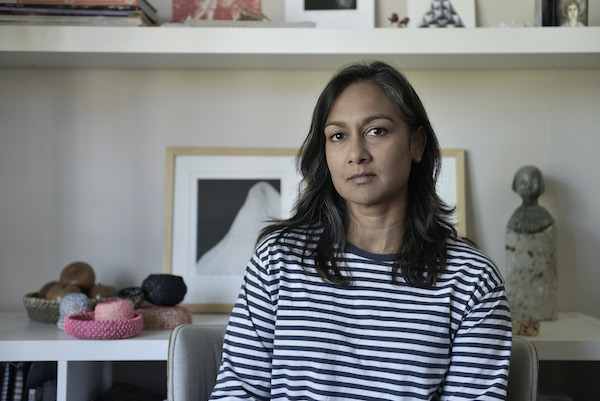 Shanti Gelmi sits in what appears to be her studio - small sculptures are visible in the background. She wears a navy and white stripy t-shirt. She looks contemplative.