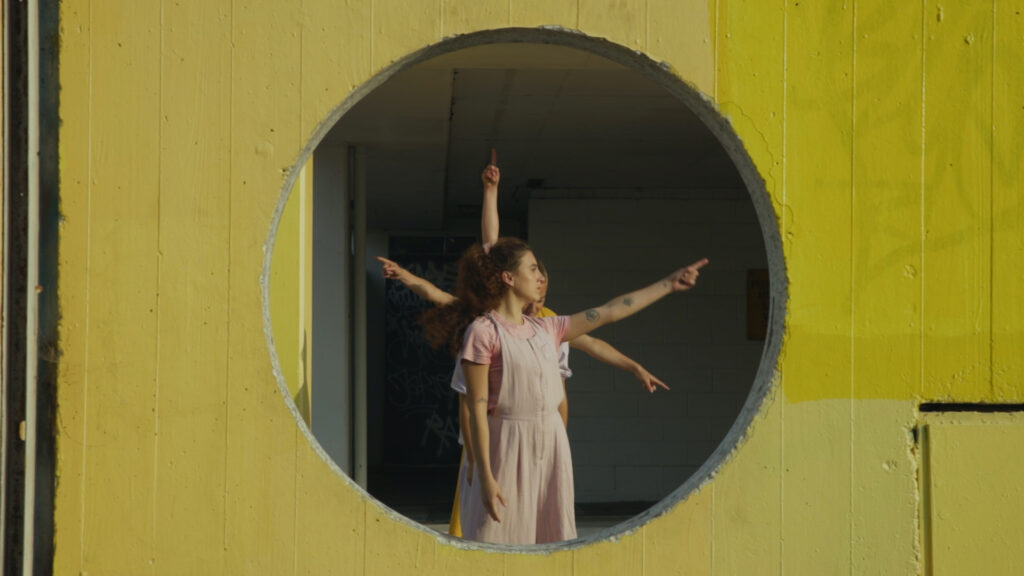 Dancers stand in a line, pointing in different directions. They are framed by a circle of concrete, painted yellow.