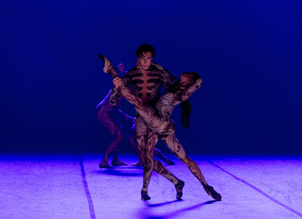 A male dancer holds a female dancer -she is in a full split and he has one hand under her extended leg and the other under her shoulders