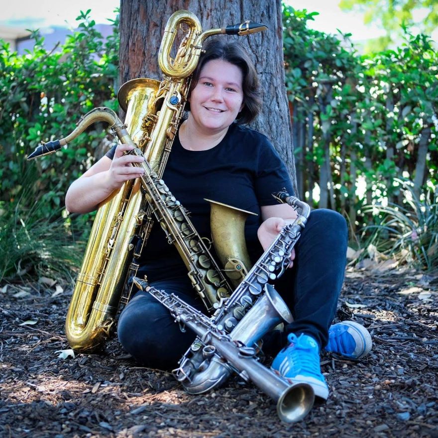 A girl leans against a tree cradling four different sized saxophones on her lap