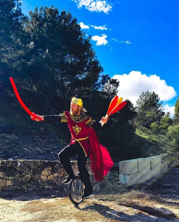 A man dressed as a medieval knight balances on a unicycle, with three skittles in one hand and a balloon sword in the other.