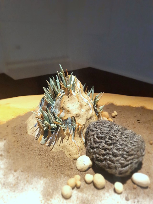 A close up of a ceramic rock, with spiky pale green protuberances, alongside a crocheted pouch, made of natural-looking fibres. These sit on a bed of sand, with small pebbles scattered about.