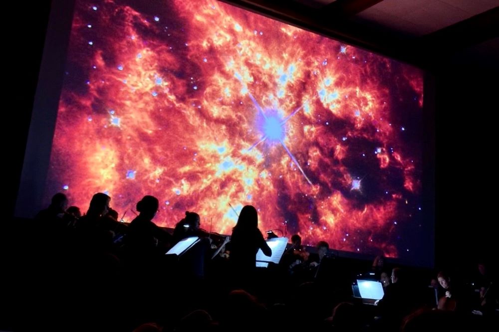 An orchestra is visible silhouetted against an big screen showing an image of a bright red solar flare