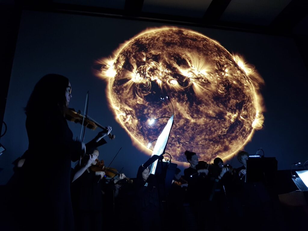 musicians stand sillouetted against a satellite photo of the sun