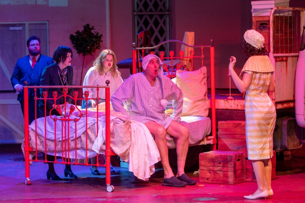 A man in a nightgown sits on a bed, surrounded by a 4 other cast members