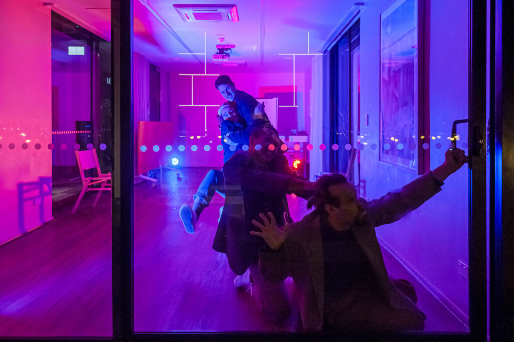 Four men are moving towards a sliding glass door. The man at the front has his faced smooshed against the glass, the others appear to be trying to stop him. The room is lit magenta.