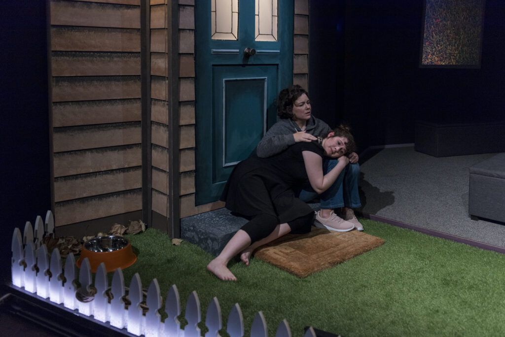 Two women sit on the front doorstep of a weatherboard house. One has her head resting on the other's lap. The house has a white picket fence and astro turf lawn.