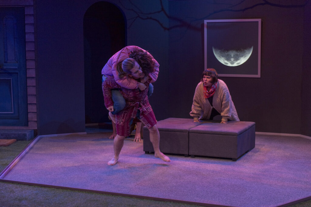 Two people appear to be wrestling in a sparsely furnished room. One has jumped on the other's back, the other appears to be trying to wrestle him off. A third person leans on a box seat, watching intently, perhaps egging them on.