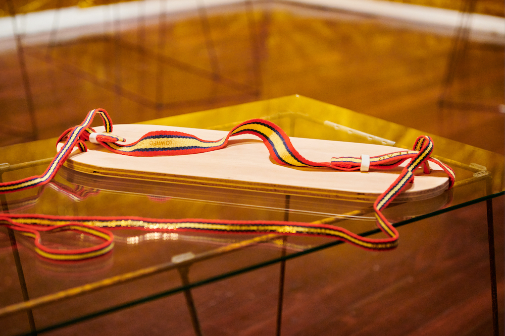 A harness made of a wooden board that is a rectangle with rounded edges, and red, navy and yellow striped strapping. The harness sits on a glass-topped table.