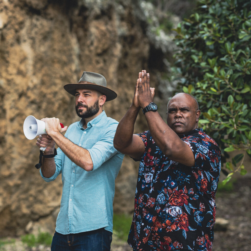 A white man and a First Nations man stand in front of a rock face, some of which is covered with vegetation. The First Nations man wears a colourful shirt and has his hands raised as though he has just clapped. The white man wears a hat and a turquoise shirt and holds a small megaphone.