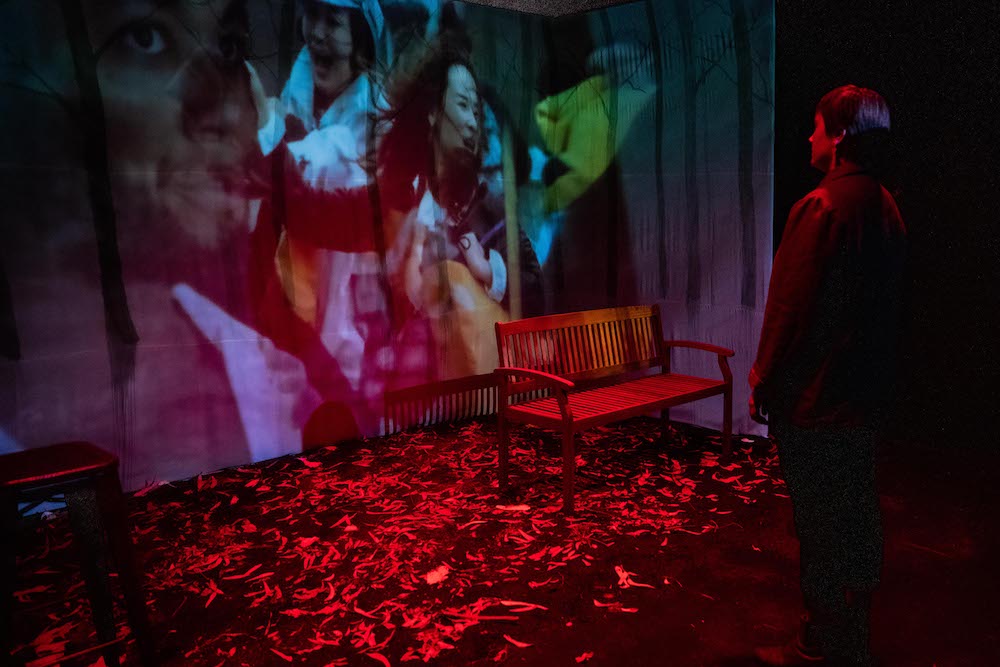 Katie McAllister stands on stage, gazing at a projection that is a collage of people who seem to be screaming. In front of the projection are dead leaves, a park bench and a stool.