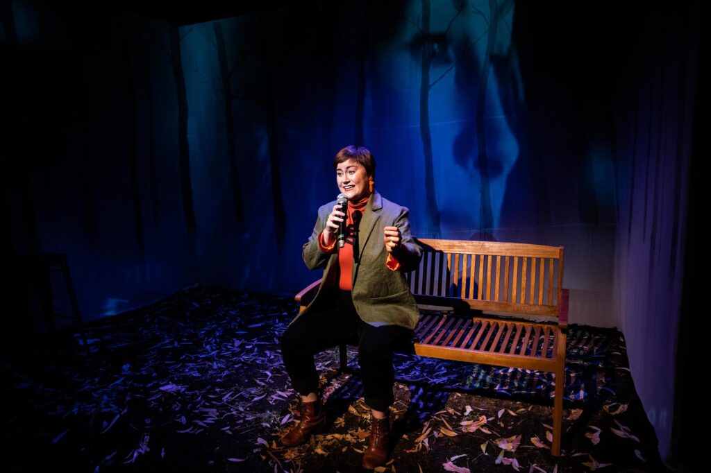 A young woman sits on a wooden park bench. She wears a khaki blazer over a red skivvy and black pants. The floor is scattered with dead leaves and silhouettes of slim gum trees are projected against the deep blue of an early night sky.
