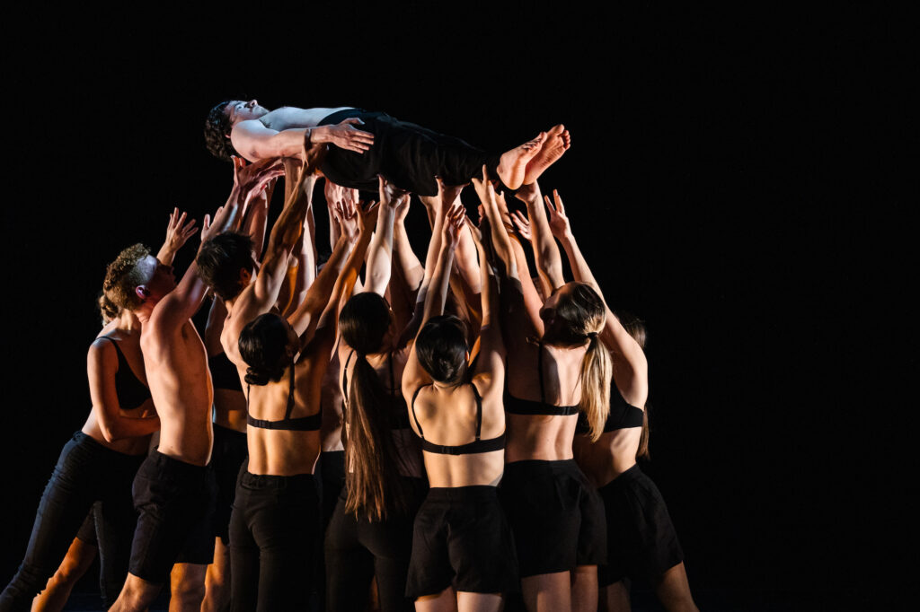 One dancer is lifted by many others. All wear black pants and the women wear black bras