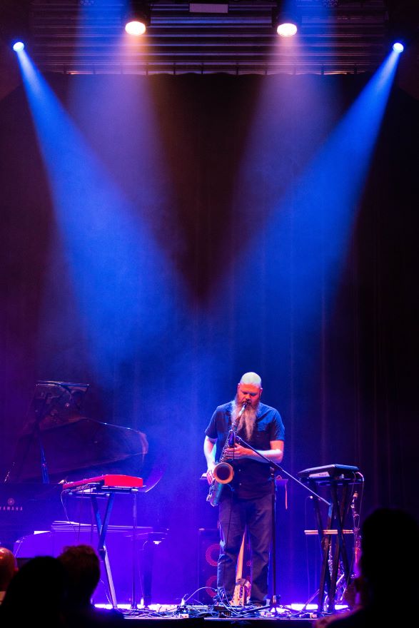 A man with long white beard and bald head stands under two spotlights while playing saxophone