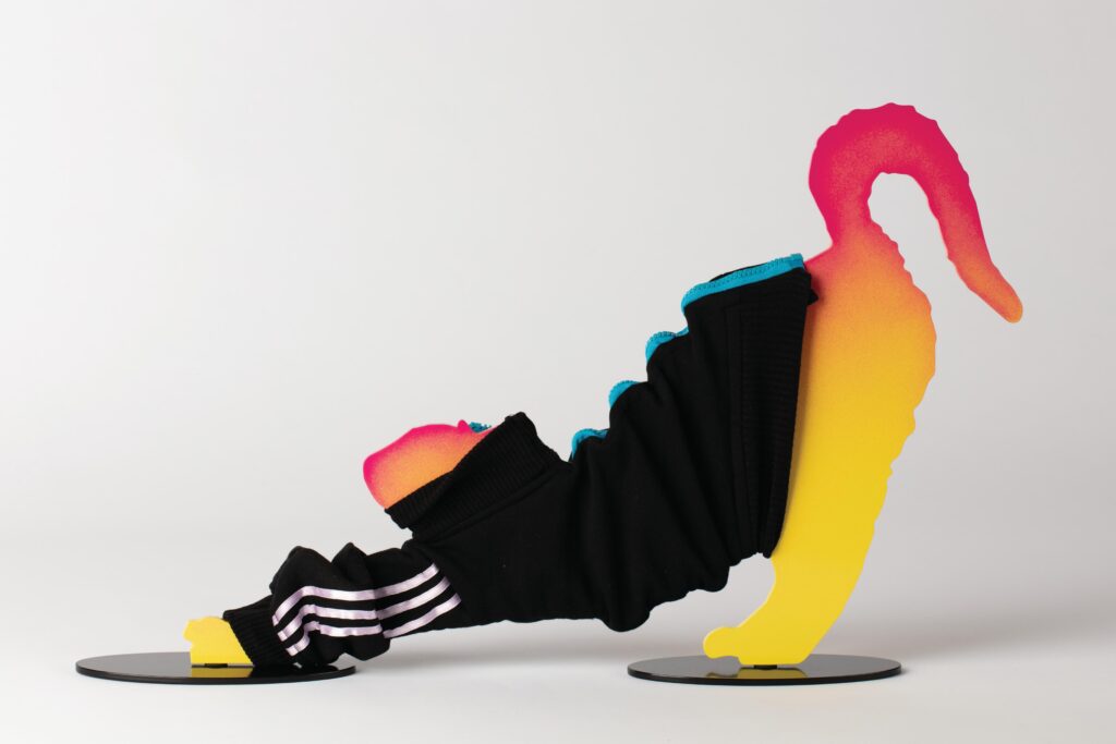 A 2-D sculpture of a cat stretching in the downward dog position. The cat is neon pink and bright yellow and wears a black Adidas-style sweater with white stripes on the forearms