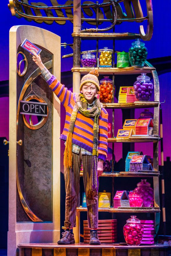 A boy wearing a scarf and beanie stands in a sweet shop holding aloft a bar of chocolate