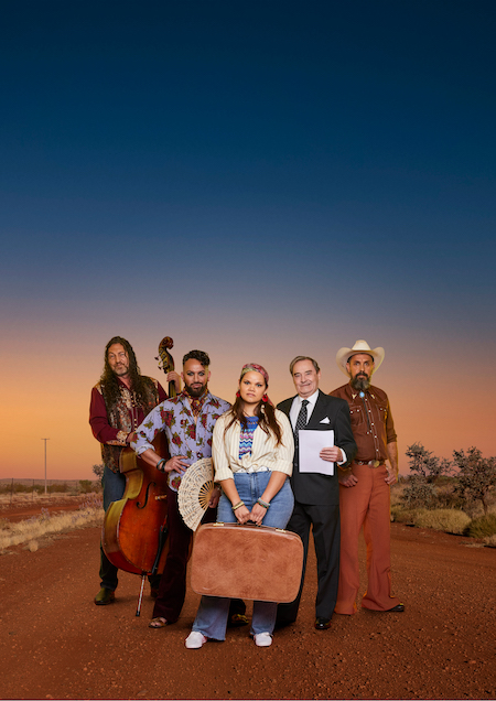 A group of 5 people stand on a red dirt road through the desert, dressed in 60s fashion. Four are First Nations people, one is white.