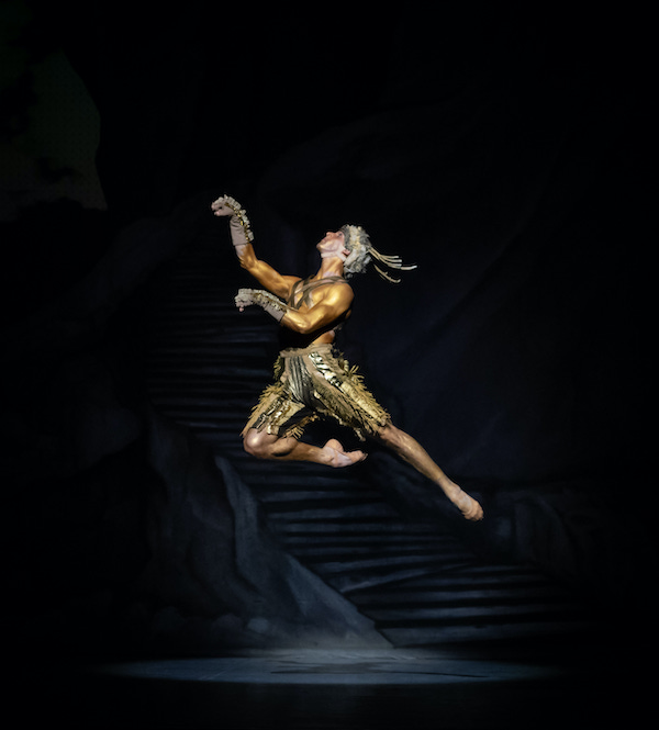 A male dancer in a golden leotard leaps in the air. He holds his hands like an animal's hooves.