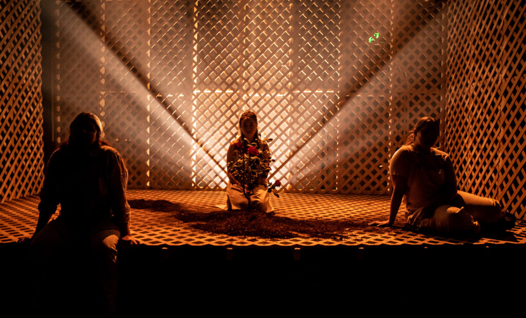 Three women sit inside a latticed box; two downstage, one centre back, in a triangle formation. The woman at the back holds flowers and behind her beams of light radiate outward and upwards, so that their faces are cast into shadow.