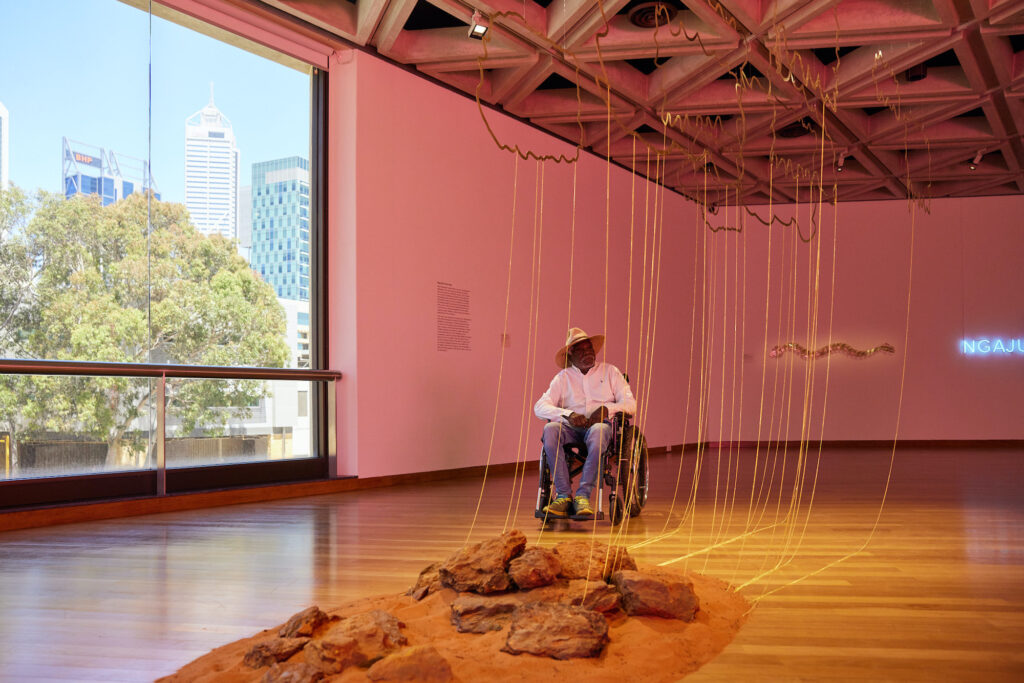 Artist Tommy May sits in a wheelchair behind his work which includes wire outlines of golden clouds and trails of gold chains that end in a pile of red dirt and rocks.