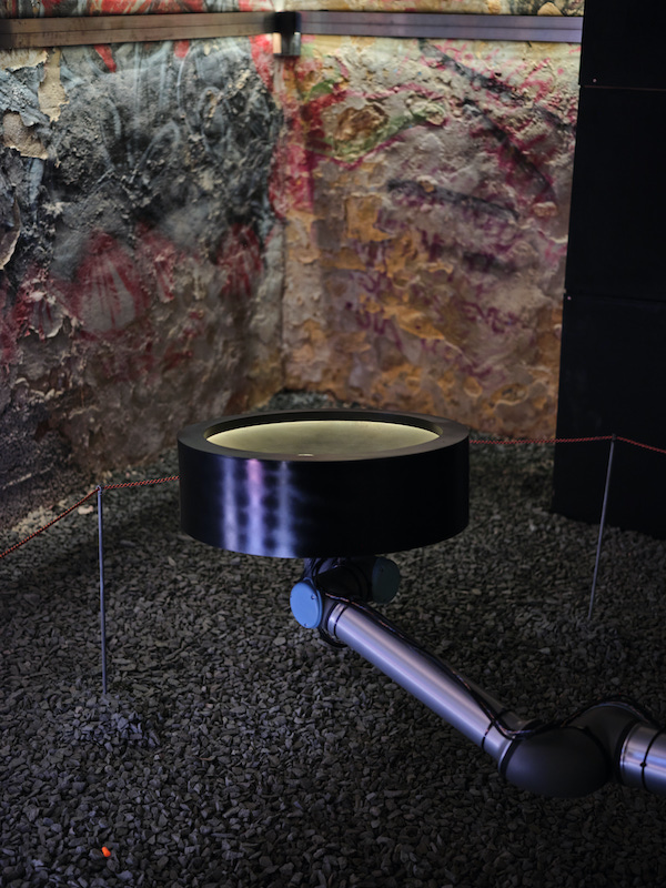 A metal arm with a disc on the end extends in front of a limestone wall that is decorated with what almost looks like colourful graffiti. The floor is gravel.