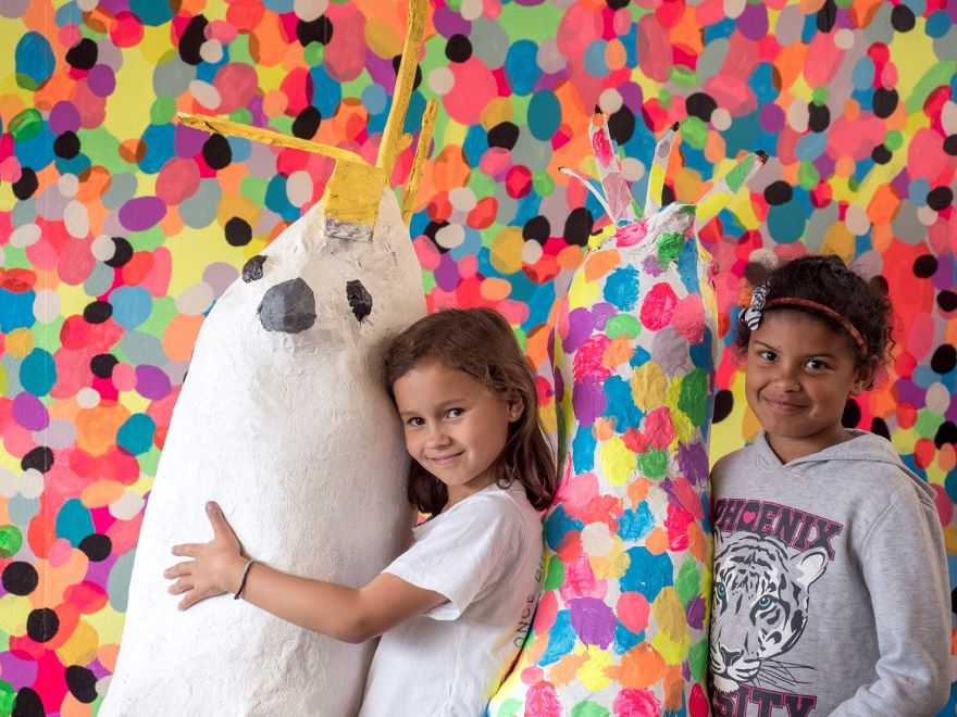 Two children hug life-size paper mache  animals in front of a wall covered in small bright dots