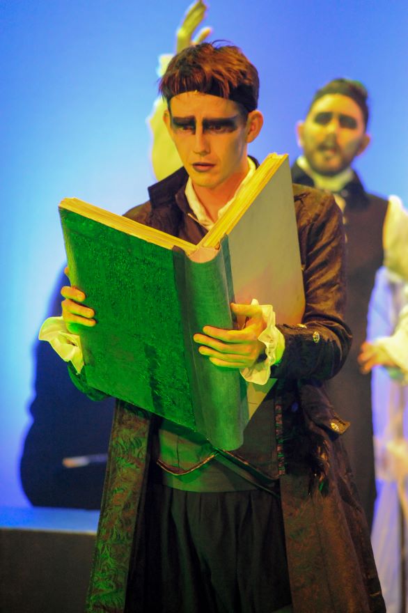 A man with black eye make up stands holding an enormous open book