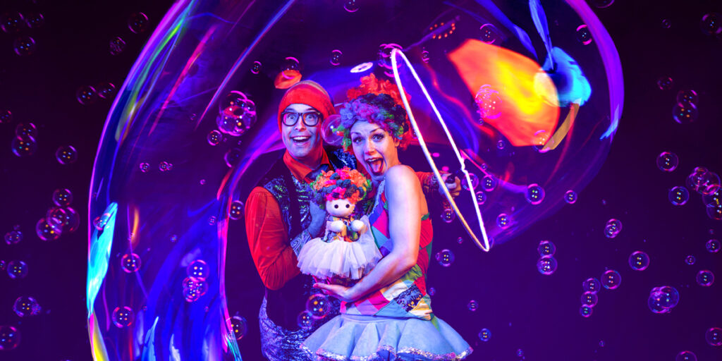 A man and woman wearing red clown wigs and holding a small puppet are surrounded by bubbles