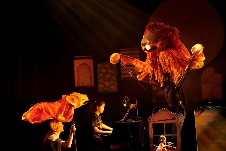 A pianist sits at her instrument on a stage, with large orange puppets oon sticks overshadowing her