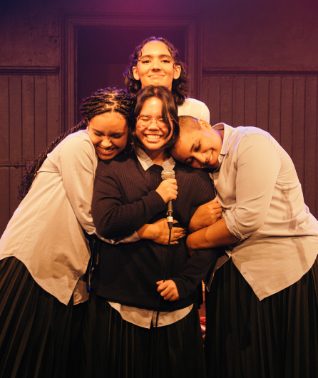 A snapshot from the musical 107. Pictured is four actors smiling as they embrace each other.
