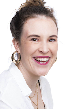 A head shot of Isablla Boladeras. She wears a white shirt against a white background, bright pink lipstick and gold hoop earrings. She is grinning at the camera.