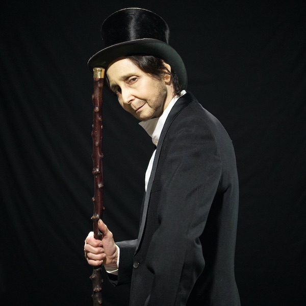 Pictured for Fringe World's show '2 Marys' is a woman dressed as a man. She has light stubble and wears a black top hat and suit jacket over a white shirt. She holds what looks like a cane in front of her which she uses to lift her hat slightly.