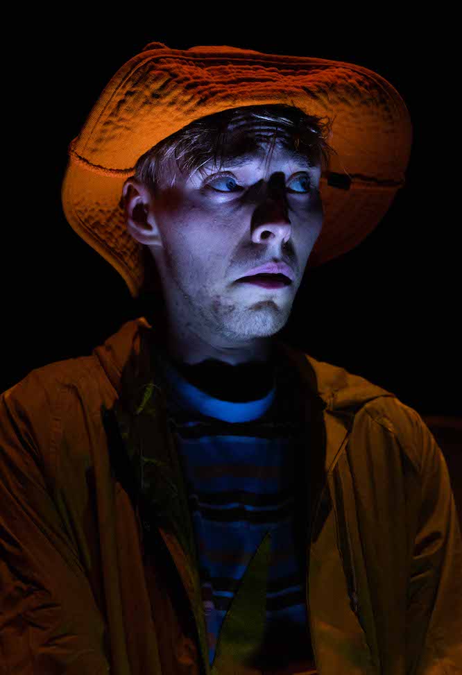 Alex from Alex and Evie and the Forever Falling Rain. Pictured is a close up of a young man in a darkened room with torchlight shone on his face from below. He is wearing a raincoat and hat and looks worried.