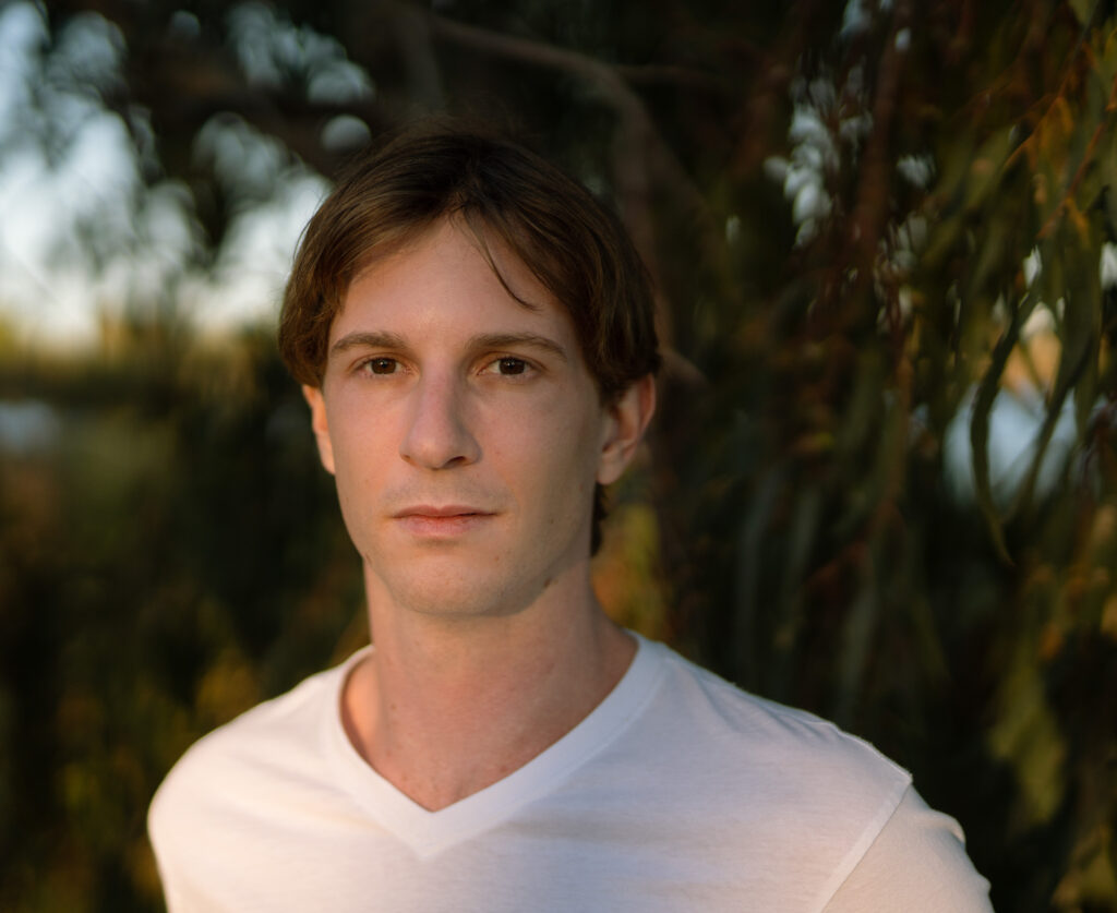 The headshot of Salome creator Andrew Sutherland. Pictured you see his shoulders up, he is wearing a white shirt in front of a vegetation background.